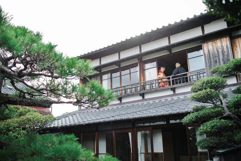 どうだん亭の外観と手入れされた松。2階の縁側に立つ和装の新郎新婦を遠景で切り取った前撮り