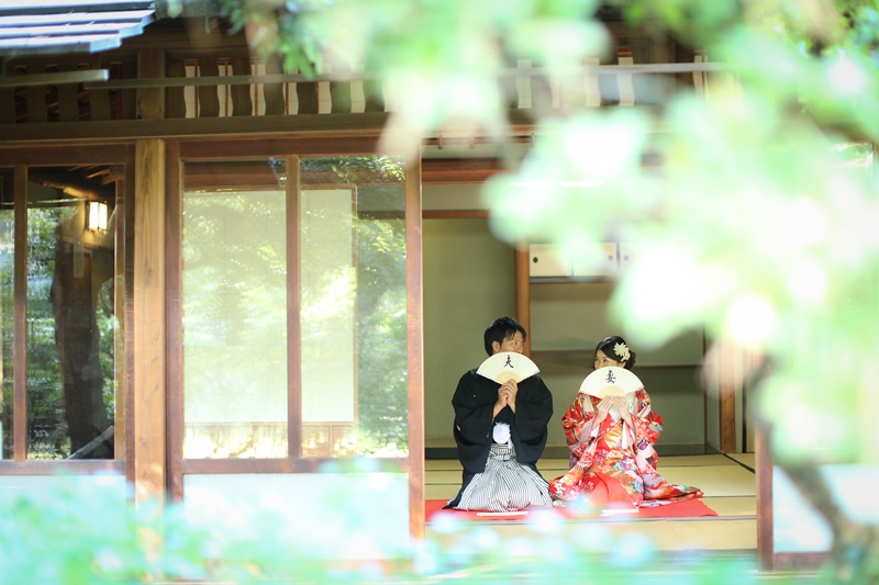 新緑越しに茶室の縁側で並んで座る色打掛の花嫁と紋付袴の新郎。木漏れ日がやわらかい和装前撮り