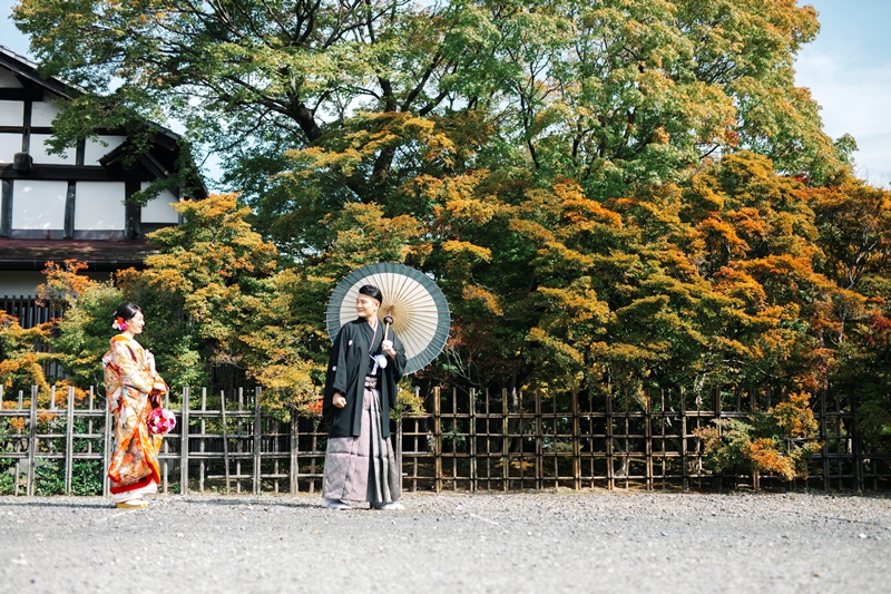 色づいたどうだんつつじを背に和傘を差して立つ色打掛の花嫁と黒紋付袴の新郎。秋の和装前撮り