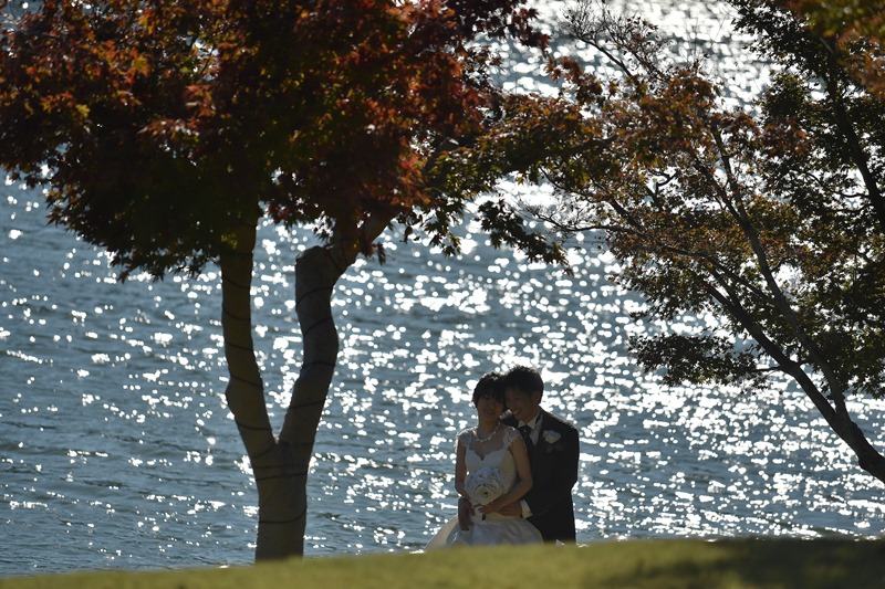きらめく湖面と色づいた木を背に寄り添うウェディングドレスの花嫁と新郎。秋のロケーションフォト