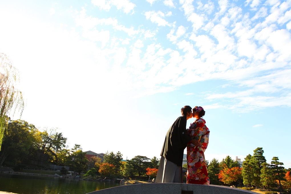 龍仙湖のほとりで青空を背に寄り添う色打掛の花嫁と紋付袴の新郎。水辺のロケーションフォト