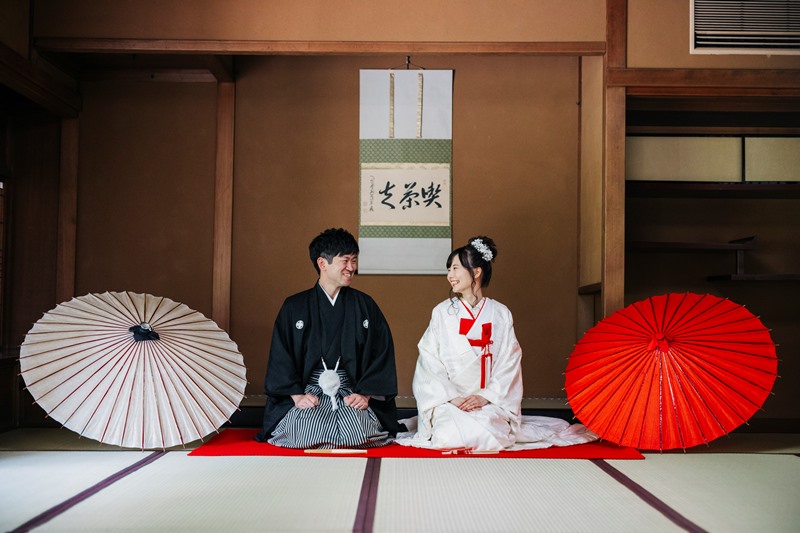 赤と白の和傘を広げた和室で正座する白無垢の花嫁と紋付袴の新郎。掛け軸を背にした和装前撮り
