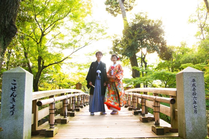 木の橋を並んで渡る色打掛の花嫁と黒紋付袴の新郎。緑に包まれた庭園で撮る和装前撮り