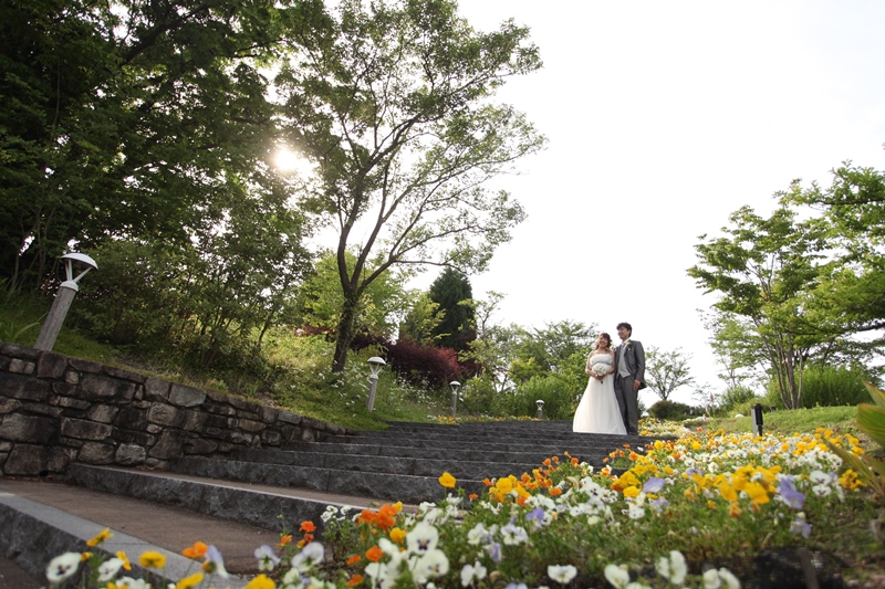 色とりどりの花壇と夕方の光に包まれて歩く新郎新婦。モリコロパークで叶えるフォトウェディング