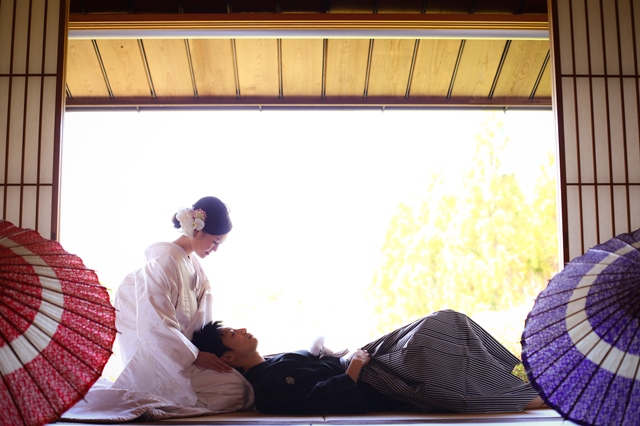 赤と紫の和傘を置いた明るい和室で、膝枕をする色打掛の花嫁と黒紋付袴の新郎。遊び心のある和装フォトウェディング