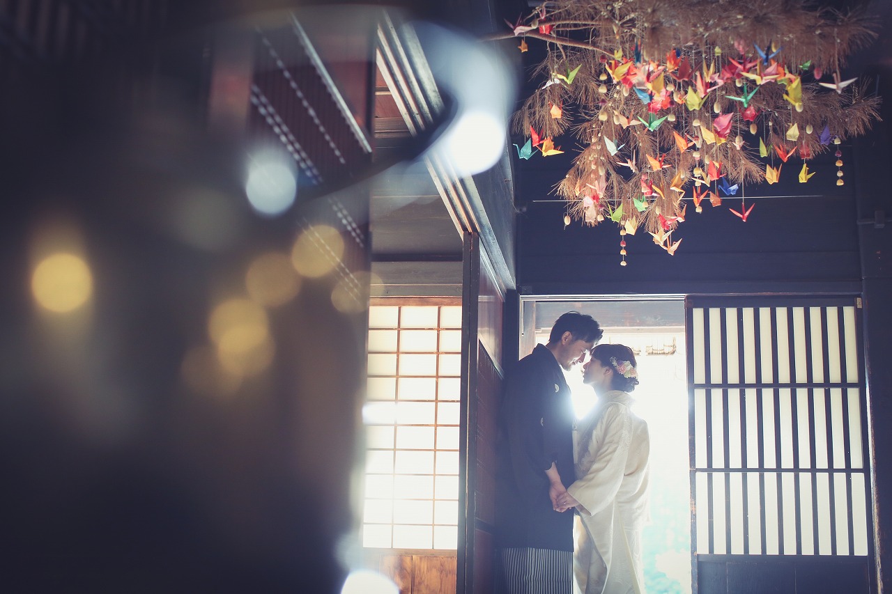 天井から折り鶴が下がる和室で、障子の光を受けて寄り添う白無垢の花嫁と紋付袴の新郎。逆光が美しい和装前撮り
