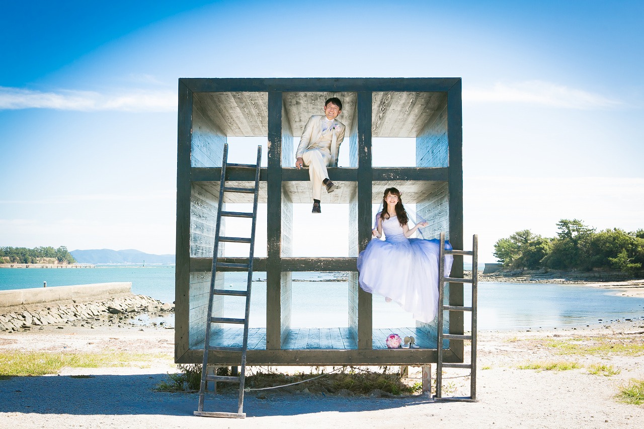 佐久島のおひるねハウスの枠の中に座る新郎とドレス姿の花嫁。海と空に囲まれたアートスポットでのフォトウェディング