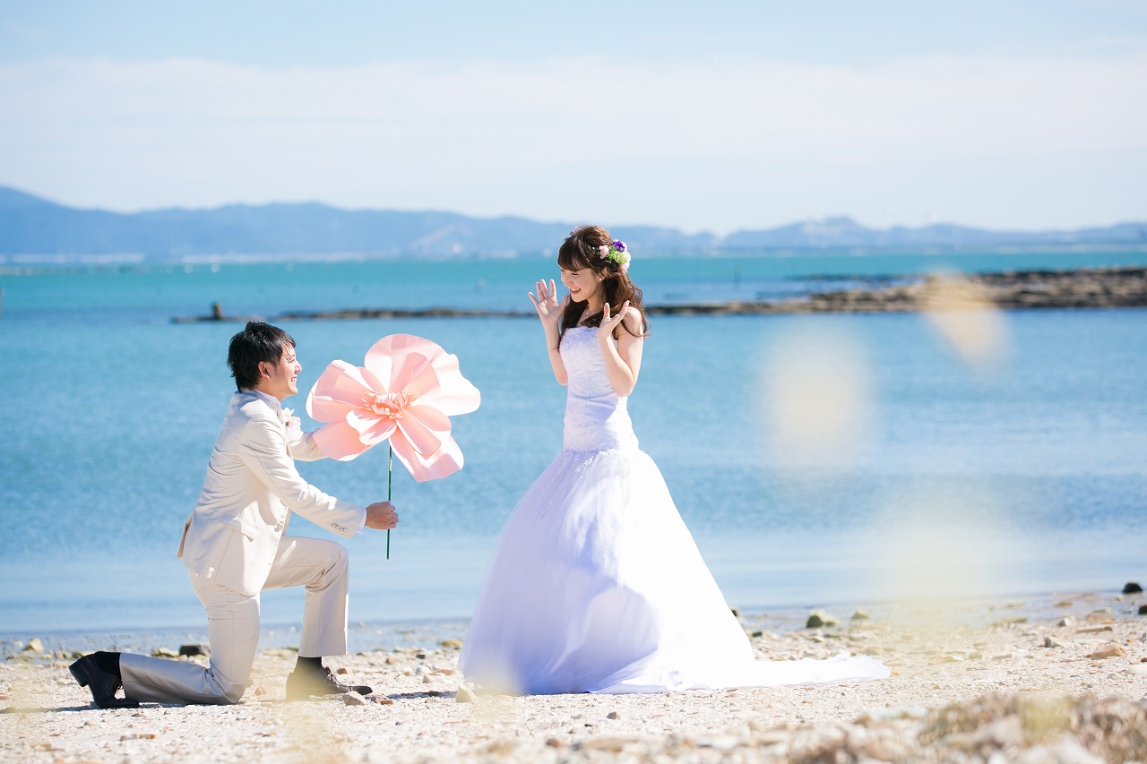 穏やかな海辺で大きな花のアイテムを持って向かい合う新郎新婦。青い海と空が広がる洋装ロケーションフォト