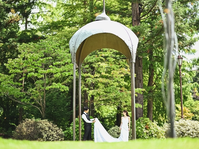 白いガゼボの下でトレーンを広げる花嫁と手を取る新郎。緑に囲まれたフォトウェディング