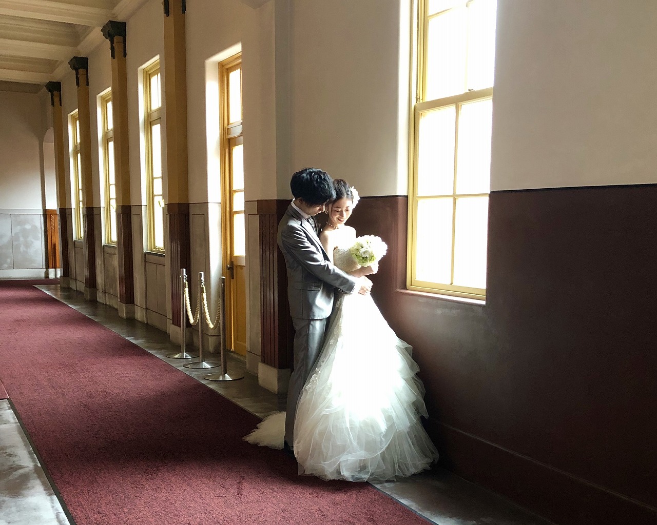 赤い絨毯の廊下で窓の光を受けて寄り添う花嫁と新郎。マーメイドドレスが映えるクラシカルな前撮り