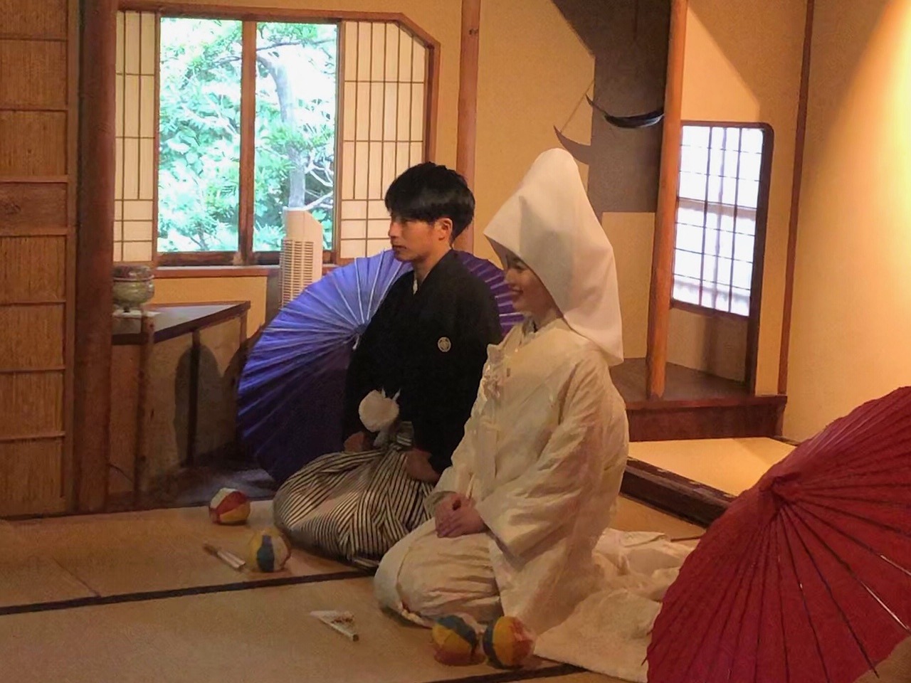 和室で赤い和傘を広げて正座する白無垢の花嫁と紋付袴の新郎。落ち着いた雰囲気の和装フォトウェディング
