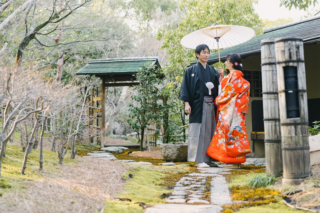 苔むした飛び石の先に立つ色打掛の花嫁と紋付袴の新郎。和傘を差して歩く静かな庭のフォトウェディング