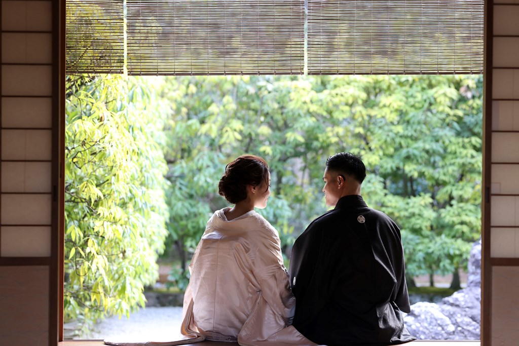 簾の下がる縁側に腰を下ろして庭を眺める白無垢の花嫁と黒紋付袴の新郎。落ち着いた和装前撮りの一枚