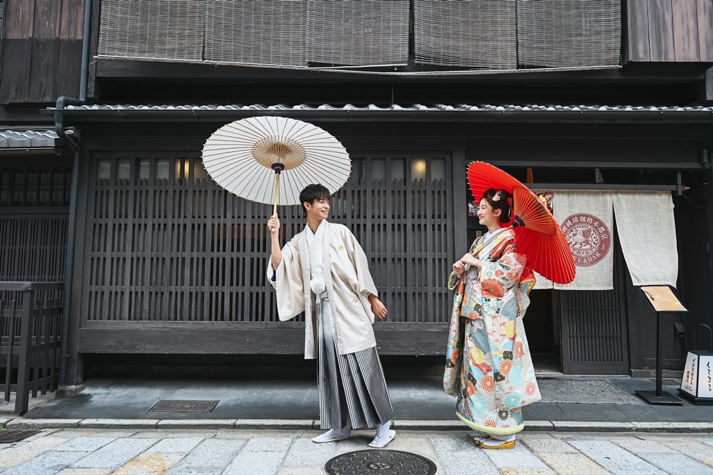 祇園の和装建築の前で和傘を差して並んで歩くふたり