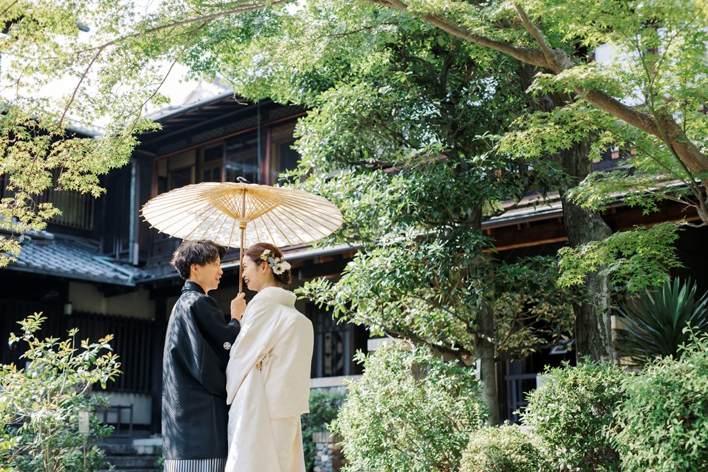 木立の中で和傘をさして向かい合う白無垢の花嫁と紋付袴の新郎。揚輝荘の緑に包まれた和装前撮り