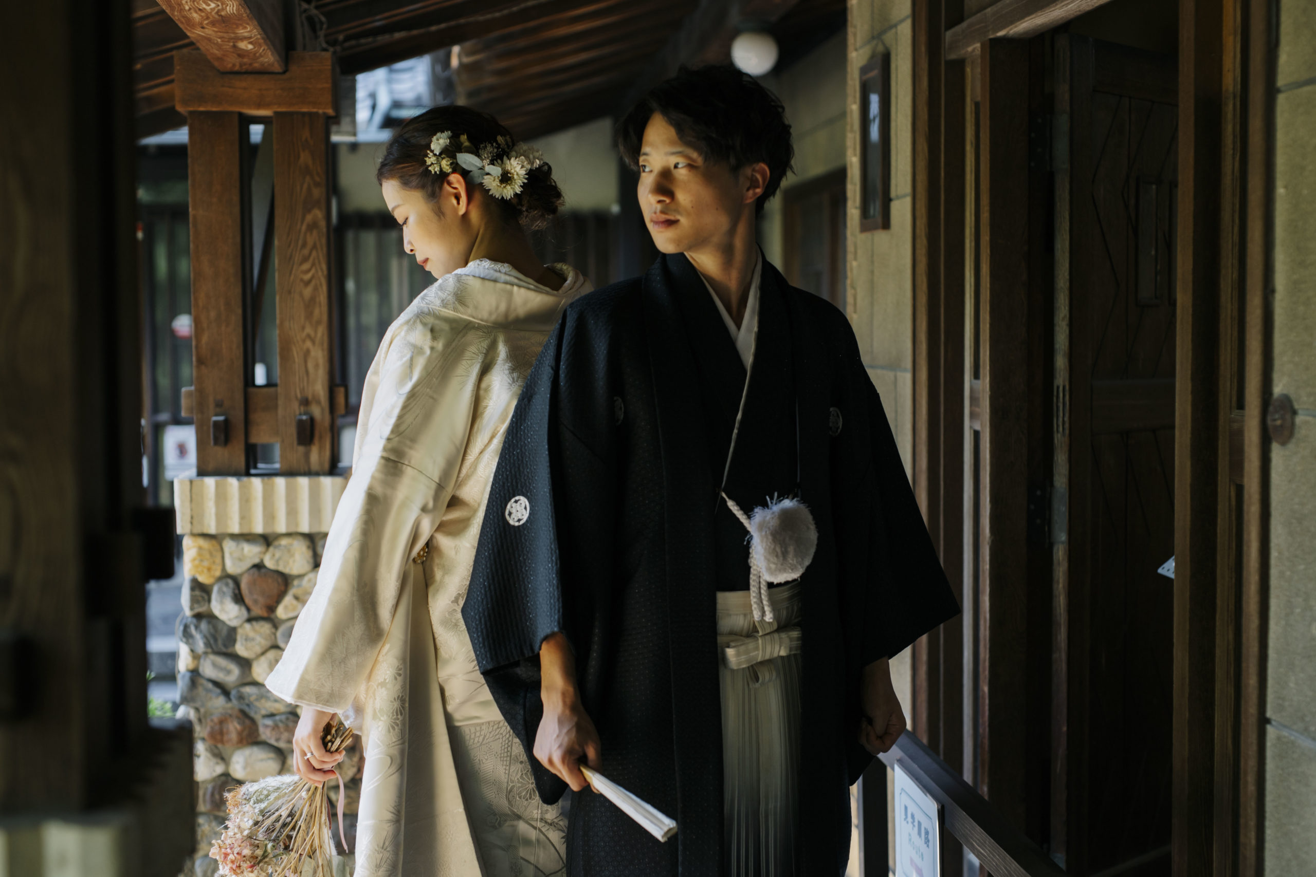 軒下で扇子を手に寄り添う紋付袴の新郎と白無垢の花嫁。落ち着いた表情が残せるロケーションフォト