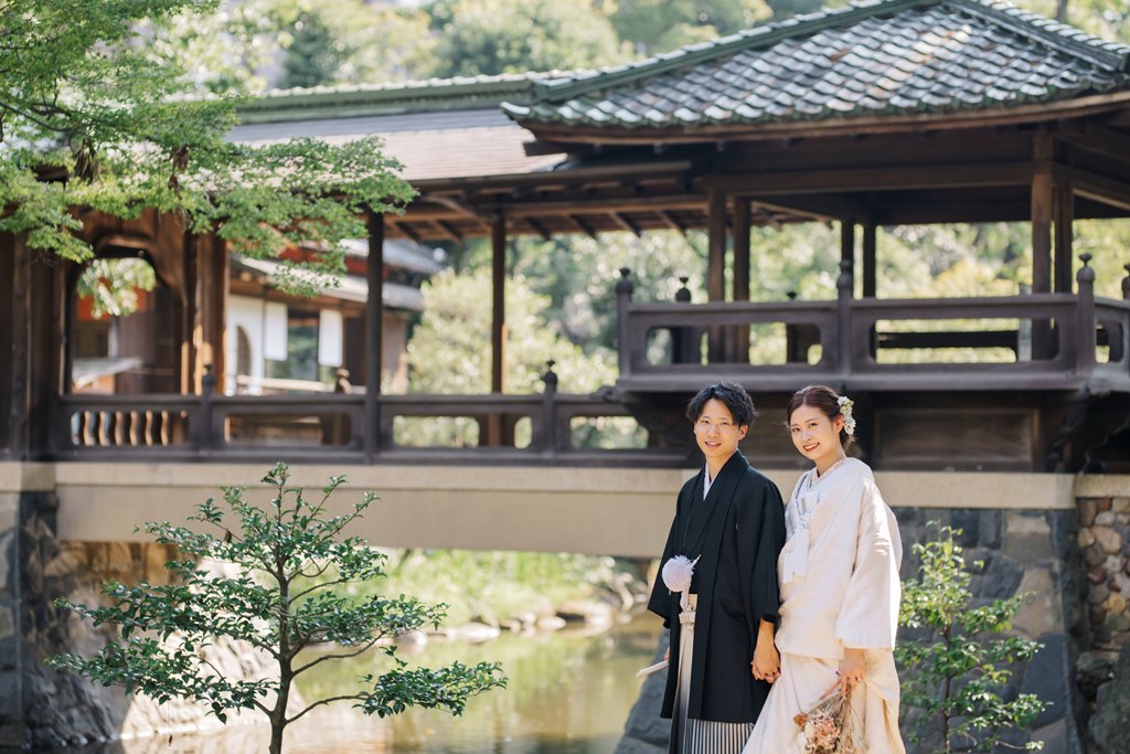 池のほとりの渡り廊下を背に立つ白無垢の花嫁と紋付袴の新郎。大正ロマンの建築が映える前撮り