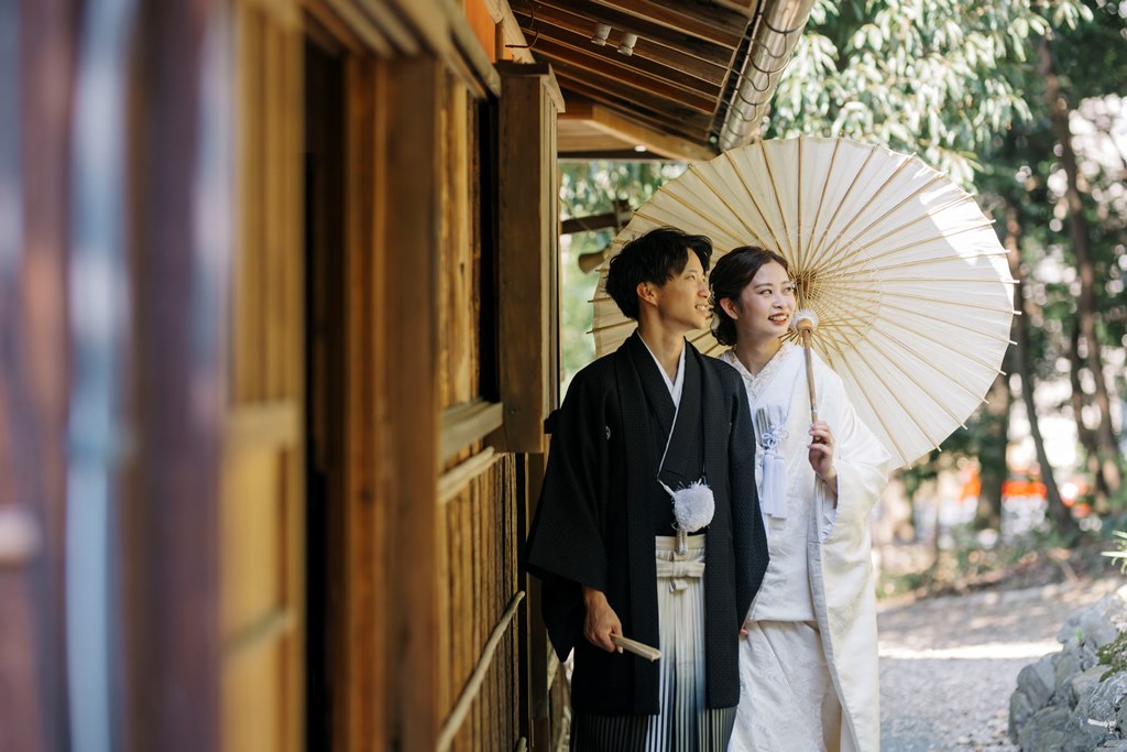 建物の縁側沿いを和傘をさして歩く白無垢の花嫁と新郎。木の質感が伝わるしっとりとした和装前撮り
