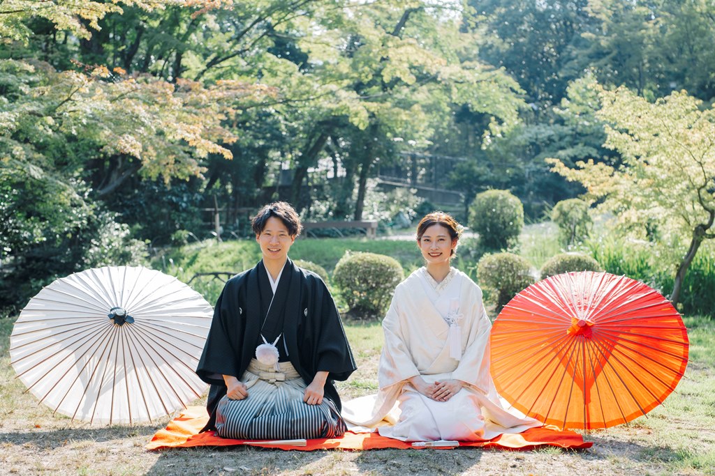 赤と白の和傘を広げて庭園で正座する、白無垢の花嫁と紋付袴の新郎。揚輝荘の和装前撮りで人気の正座のポーズ