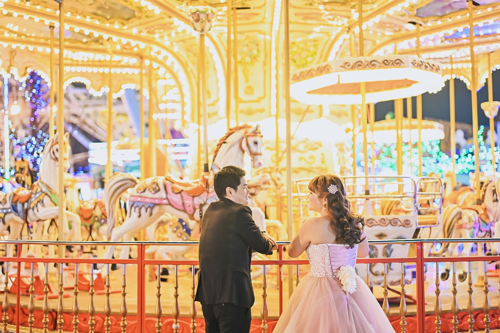 きらめくメリーゴーランドの前で見つめ合うカラードレスの花嫁と新郎。シートレインランドで撮るナイトフォトウェディング