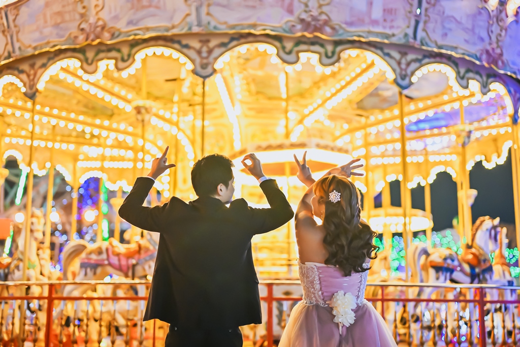 メリーゴーランドの前で両手を広げておどけるポーズをとる新郎新婦。遊園地ならではの楽しい結婚写真