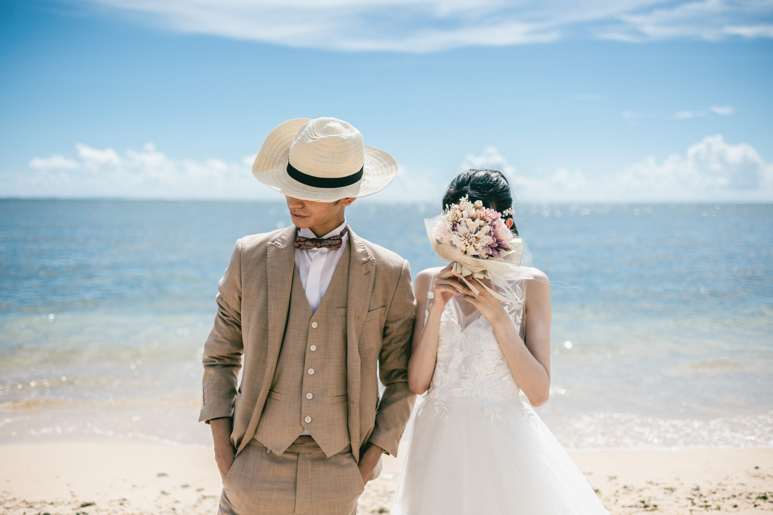 麦わら帽子とブーケを手に海を背にして並ぶ花嫁と新郎。旅の一日のようなフォトウェディング