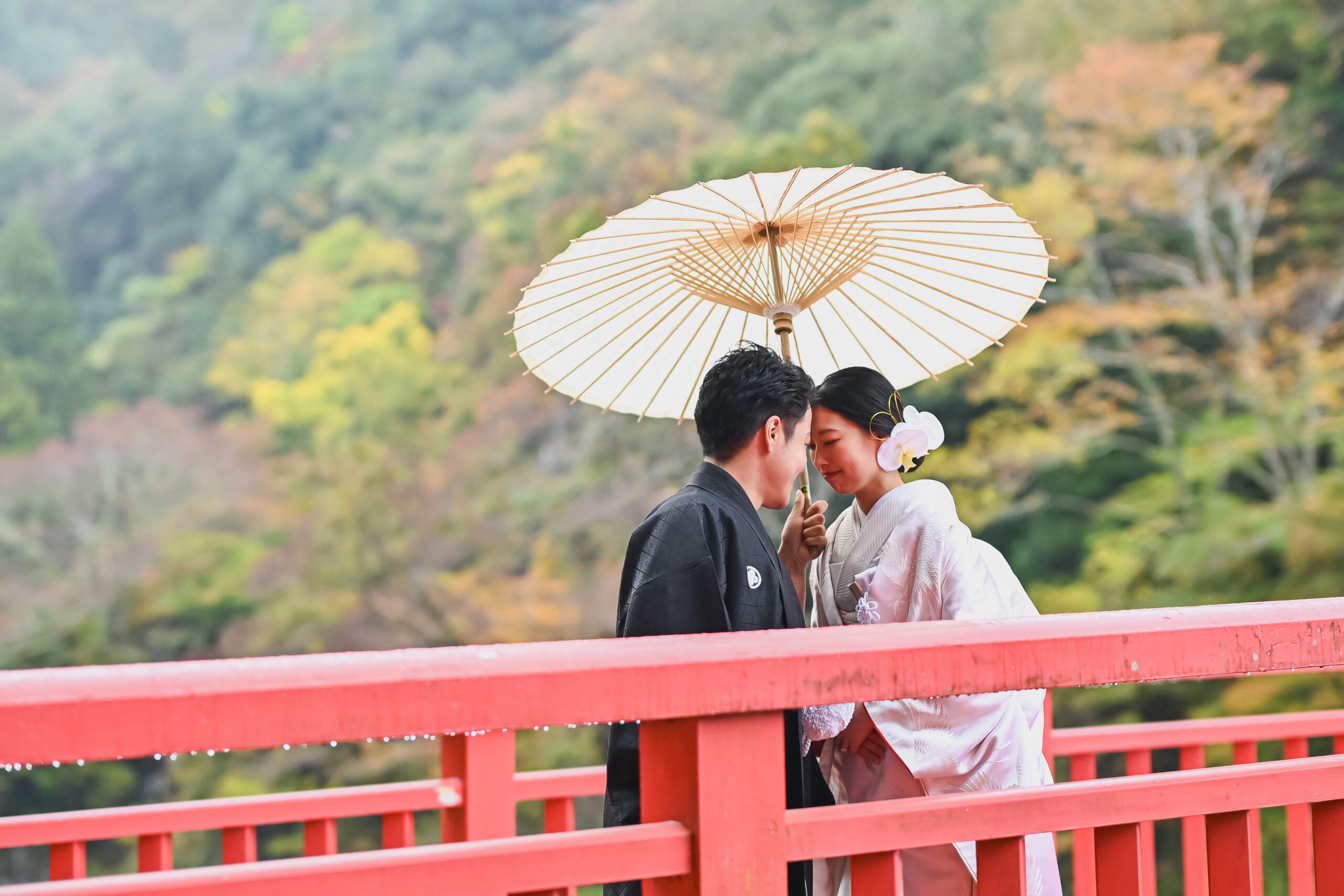 待月橋の朱色の欄干で和傘を差しおでこを寄せ合う白無垢の花嫁と紋付袴の新郎。香嵐渓で人気の前撮りポーズ