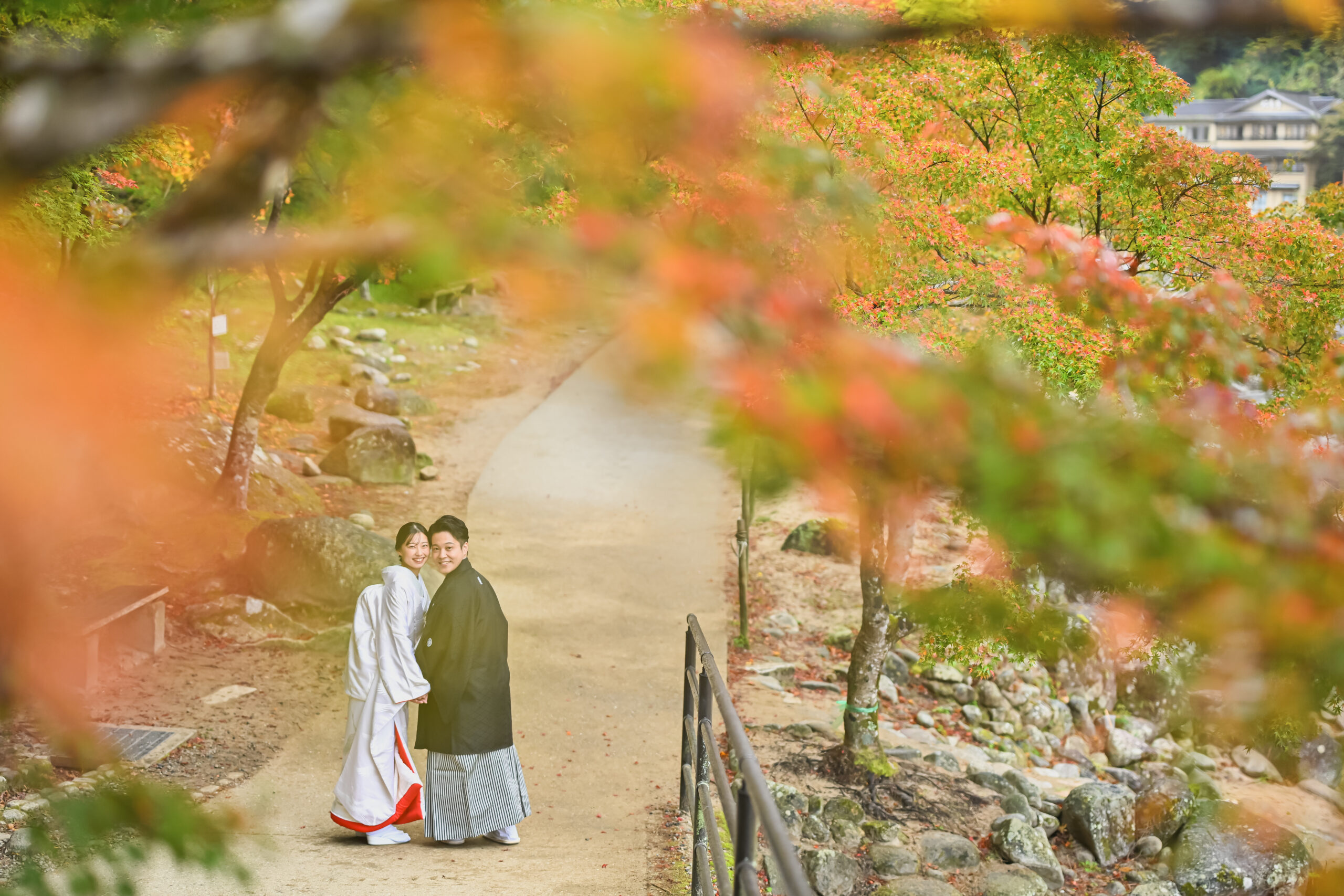 もみじ越しに遠く佇む白無垢の花嫁と紋付袴の新郎。紅葉のトンネルを生かした前撮りの構図