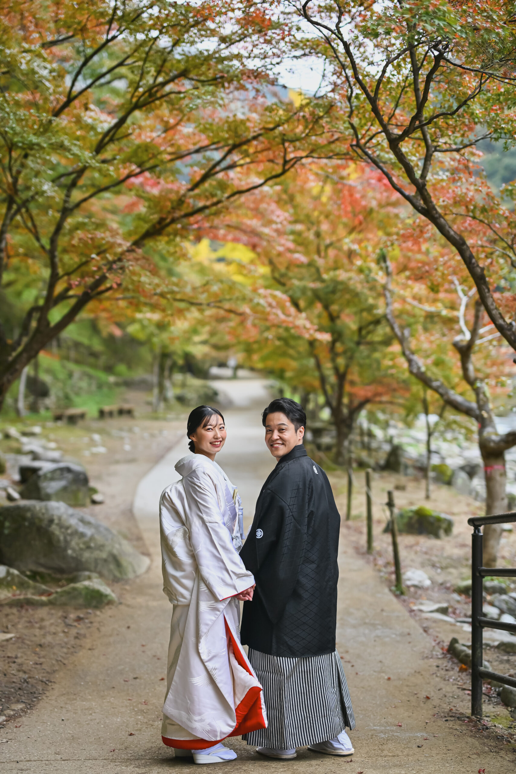 もみじが色づく遊歩道を白無垢と紋付袴で並んで歩く新郎新婦。紅葉に包まれた秋の和装前撮り