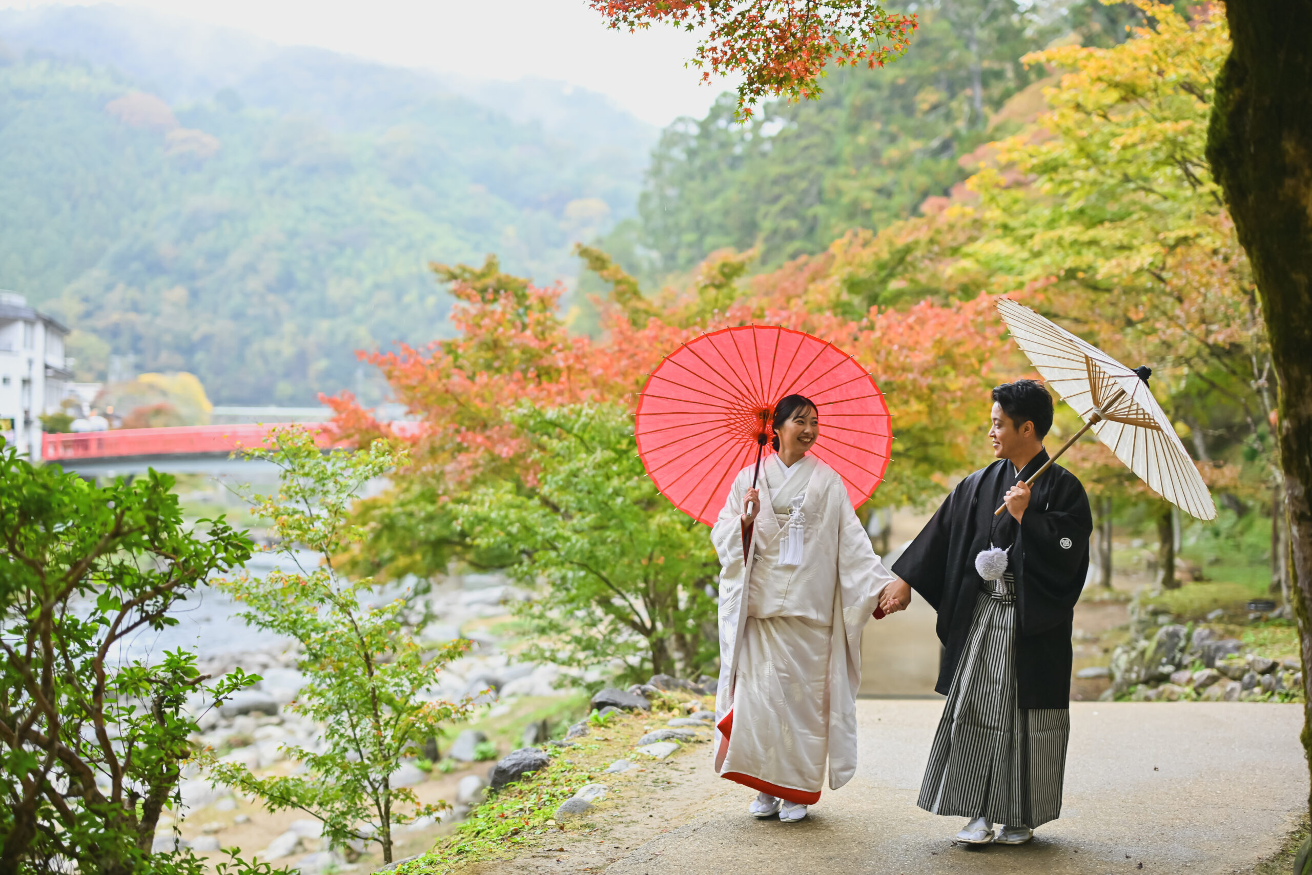待月橋と山の紅葉を背景に和傘を差して歩く白無垢の花嫁と紋付袴の新郎。香嵐渓らしい和装前撮りの一枚