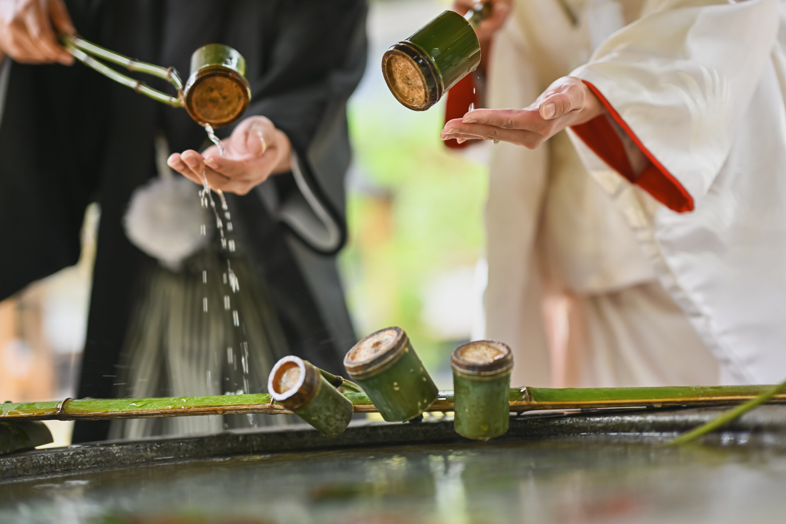 手水舎で柄杓の水を注ぎ合う白無垢の花嫁と紋付袴の新郎の手元。所作を切り取った結婚写真