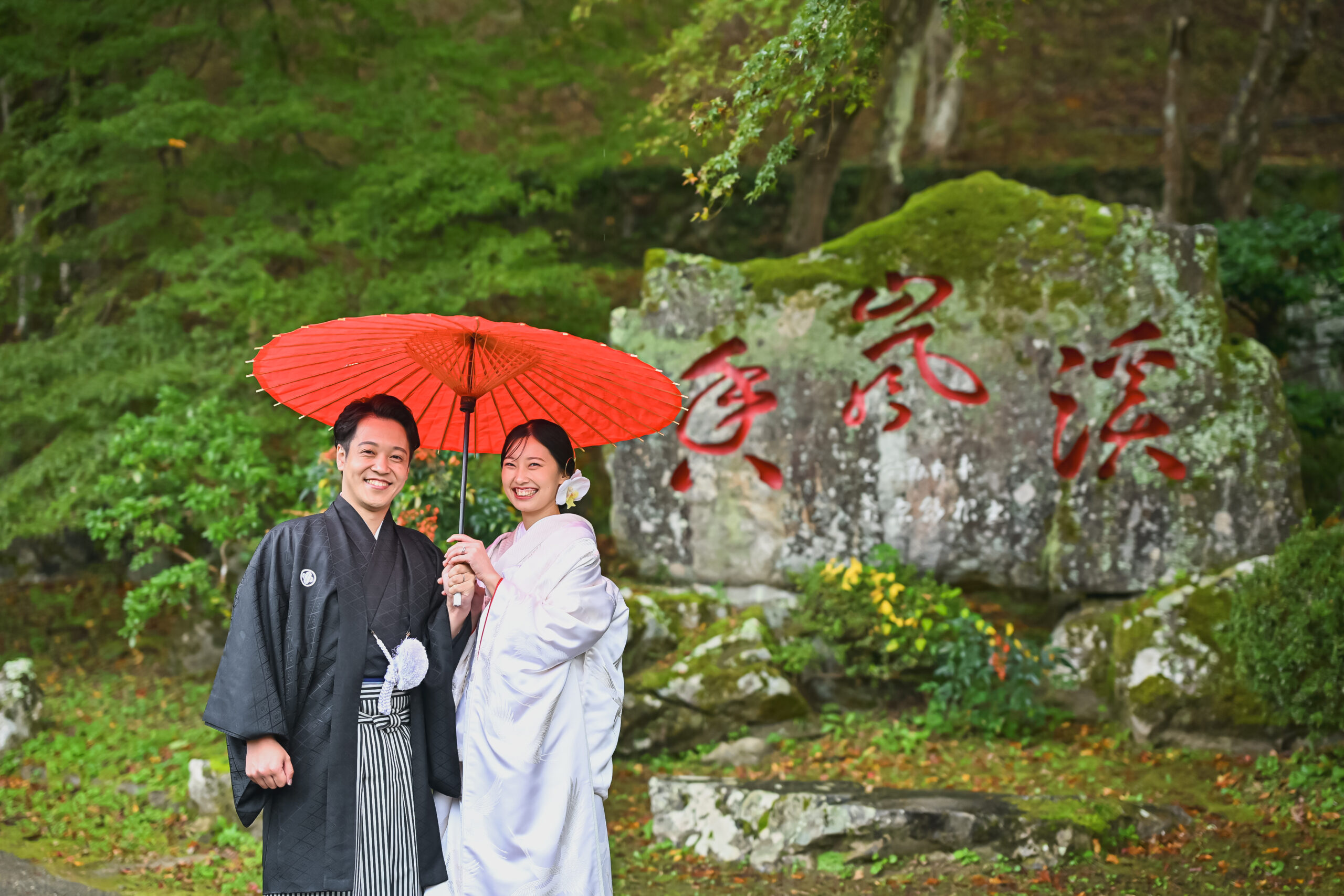 石碑の前で赤い和傘を差して寄り添う白無垢の花嫁と紋付袴の新郎。秋の思い出になるフォトウェディング