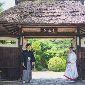 茅葺き屋根の門の下に離れて立つ黒紋付袴の新郎と白無垢の花嫁。名古屋の東山荘で撮る和装ロケーションフォト