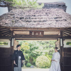 松と茅葺きの門を背に立つ黒紋付袴の新郎と白無垢の花嫁を縦位置でとらえた一枚。名古屋の東山荘で撮る和装前撮り