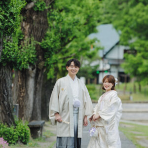 大木と茶室を背に並んで立つ白い羽織袴の新郎と白い色打掛の花嫁。岡崎公園で撮る和装ロケーションフォト