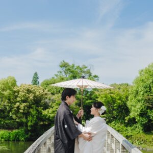 青空と緑の木々を背に、木の橋で和傘を差す黒紋付袴の新郎と白無垢の花嫁を引きでとらえた一枚。白鳥庭園で撮る和装前撮り