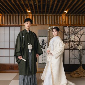 障子と梅の枝を飾った和室のセットに並んで立つ紋付袴の新郎と白無垢の花嫁の全身ショット。スタジオで撮る和装前撮り