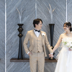 黒い花器が並ぶ石目調の壁の前で、手をつないで笑い合う蝶ネクタイの新郎と花嫁。チャペル前のロビーで撮るおしゃれな前撮り写真