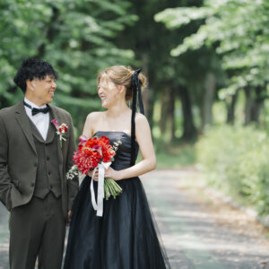 木々のトンネルの道で笑い合うオリーブのスーツの新郎と赤いブーケを持つ黒いドレスの花嫁。庄内緑地で撮る洋装前撮り