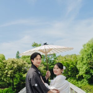 青空の下の木の橋で白い和傘を差して寄り添う黒紋付袴の新郎と白無垢の花嫁を縦位置でとらえた一枚。和装前撮りのロケーションフォト