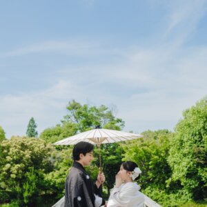 木の橋の上で白い和傘を差して見つめ合う黒紋付袴の新郎と白無垢の花嫁。日本庭園で撮る和装前撮り