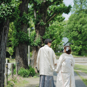 大木の並ぶ道を歩く白い羽織袴の新郎と白い色打掛の花嫁の後ろ姿を縦位置でとらえた一枚。公園で撮る和装前撮り