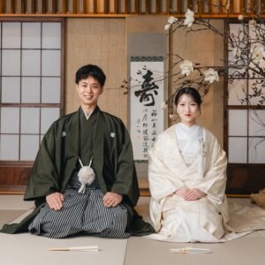 '寿'の掛け軸の前で正座して並ぶ紋付袴の新郎と白無垢の花嫁。和室のスタジオで撮った格式ある和装フォトウェディング