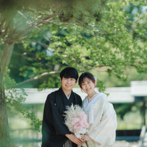 松の枝の下でピンクのブーケを持って並ぶ黒紋付袴の新郎と白い色打掛の花嫁を縦位置でとらえた一枚。日本庭園で撮る和装ロケーションフォト