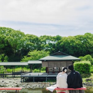 池のほとりの茶室の前で赤いベンチに座る黒紋付袴の新郎と白無垢の花嫁の後ろ姿。白鳥庭園で撮る和装前撮り