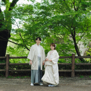 木の柵の前に並んで立つ白い羽織袴の新郎と白い色打掛の花嫁を新緑の中でとらえた一枚。公園で撮る和装ロケーションフォト