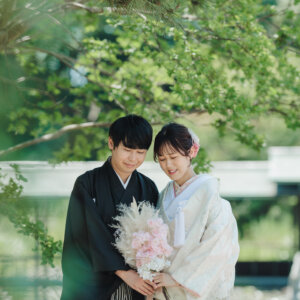 松の枝の下で寄り添い、ピンクのブーケを持つ白い色打掛の花嫁と黒紋付袴の新郎のアップ。白鳥庭園で撮る和装前撮り