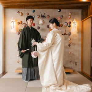 手まりと折り鶴が飾られた和室のセットで、筥迫を持つ白無垢の花嫁と扇子を手にした紋付袴の新郎。和モダンなスタジオフォト