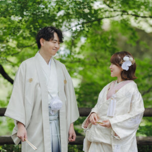 新緑の木々の下で扇子を手に見つめ合う白い羽織袴の新郎と白い色打掛の花嫁。岡崎公園で撮る和装前撮り