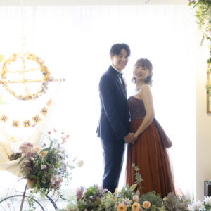 窓辺の花のアーチの前で肩を寄せて立つネイビーのタキシードの新郎とカラードレスの花嫁。やわらかな光のウェディングフォト